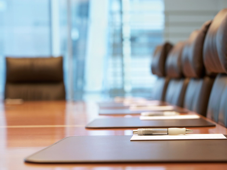 A wooden boardroom table with chairs and pens laid out on placemats, with a blurred office background.