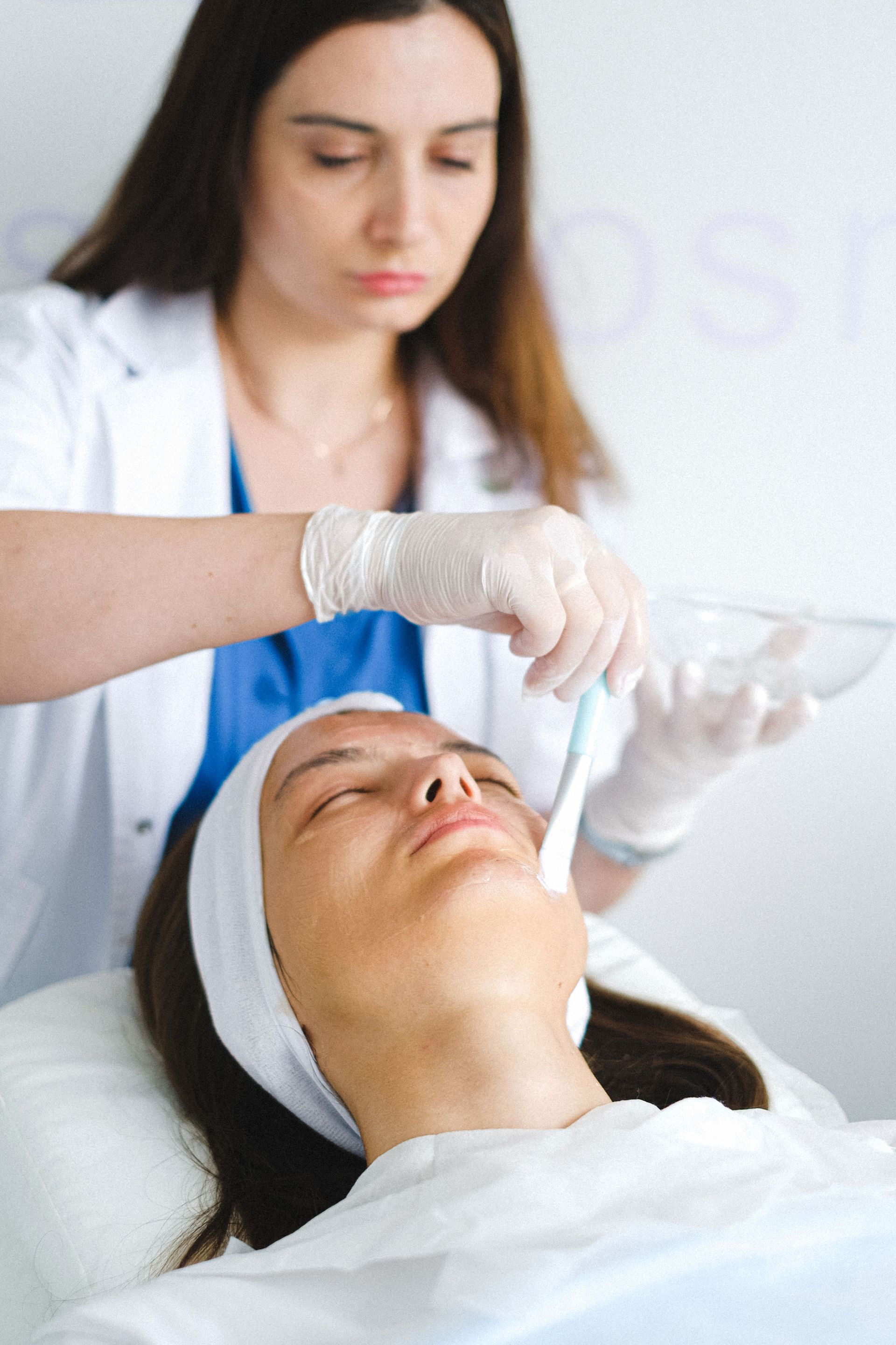 Esteticista aplicando un tratamiento a una clienta tumbada en una camilla de spa, con guantes y una diadema blanca.
