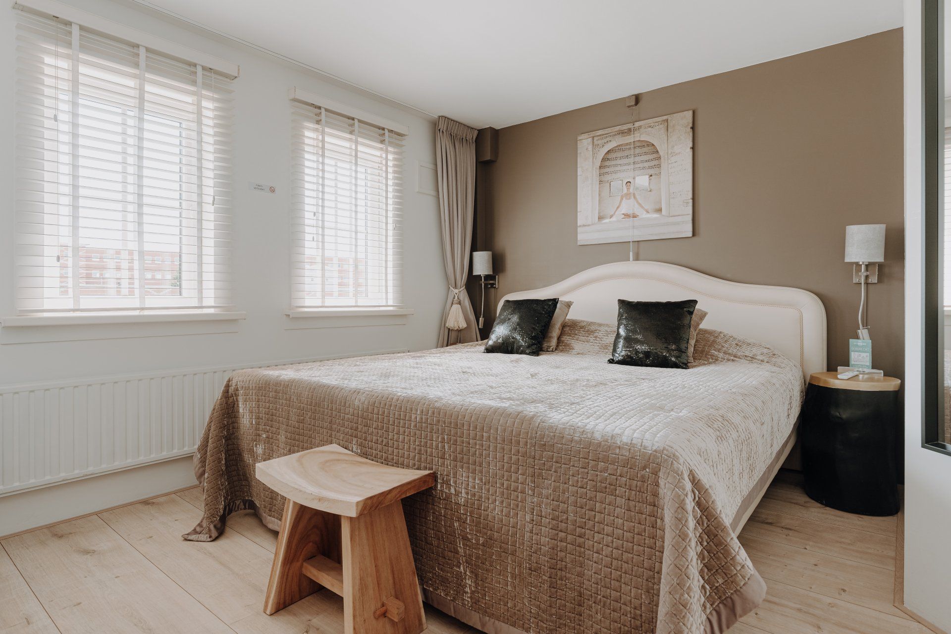 Slaapkamer met een bed, ramen met jaloezieën, een houten kruk en neutrale kleuren.