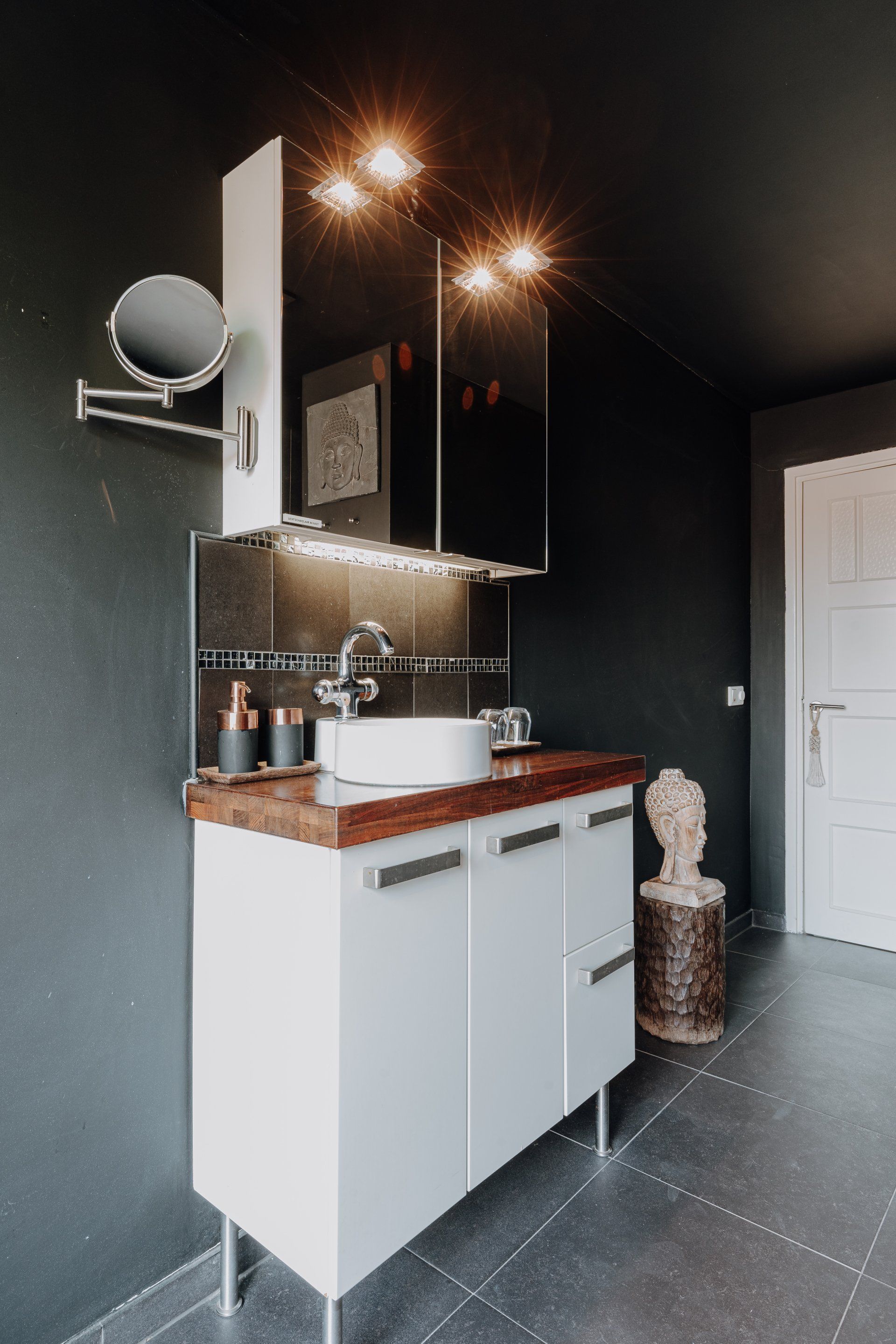 Badkamer met badmeubel, donkere muren, ronde wastafel, spiegel met verlichting en decoratieve objecten.