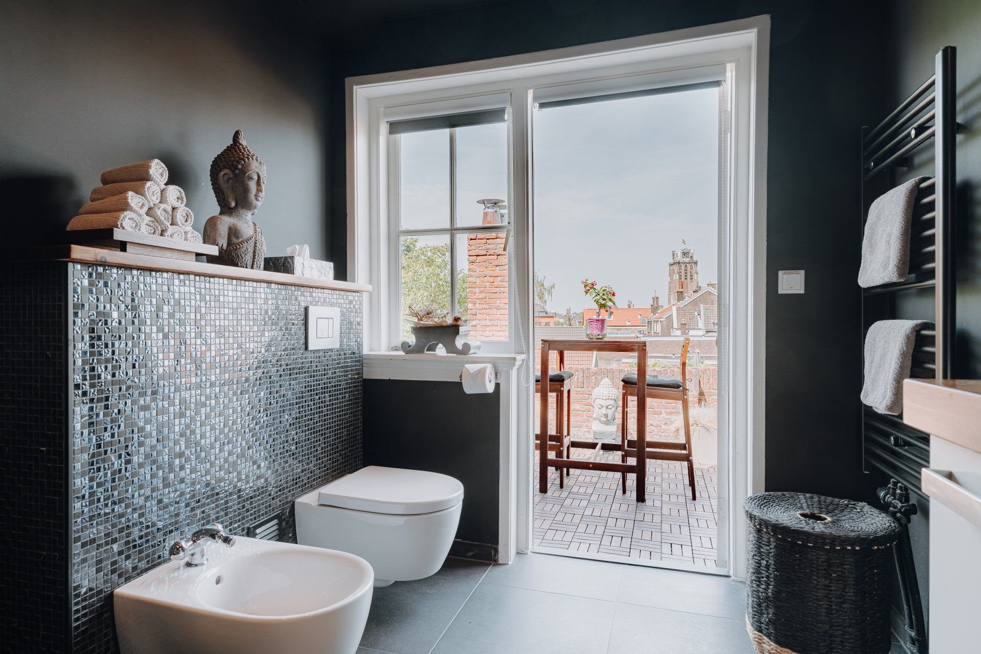 Badkamer met donkere muren, mozaïektegels, open deur naar een klein balkon met zitje en een Boeddhabeeld.