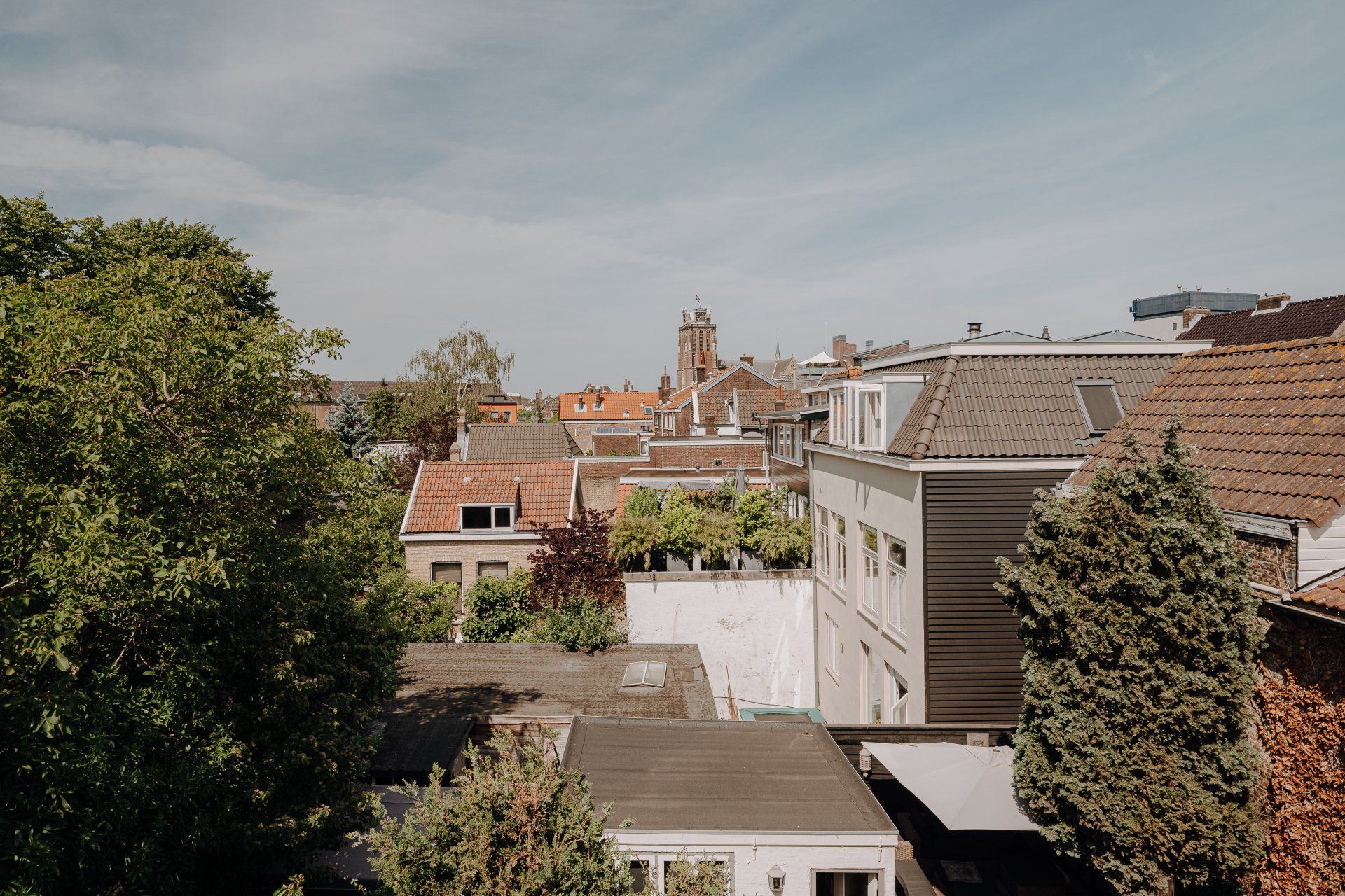 Uitzicht vanaf het dak op gebouwen met rode pannendaken, bomen en een kerktoren in de verte onder een blauwe hemel.