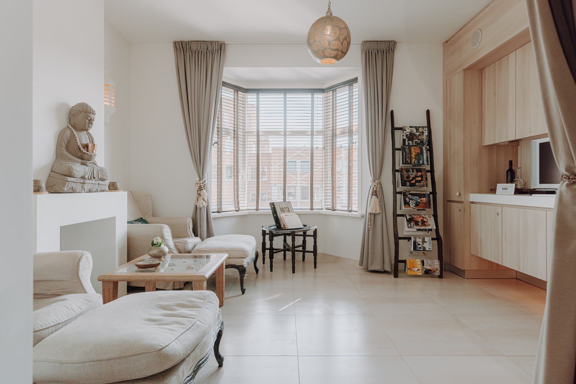 Woonkamer met Boeddhabeeld, erker, lichtgekleurd meubilair, houten jaloezieën, tv met netflix en een wandkast.