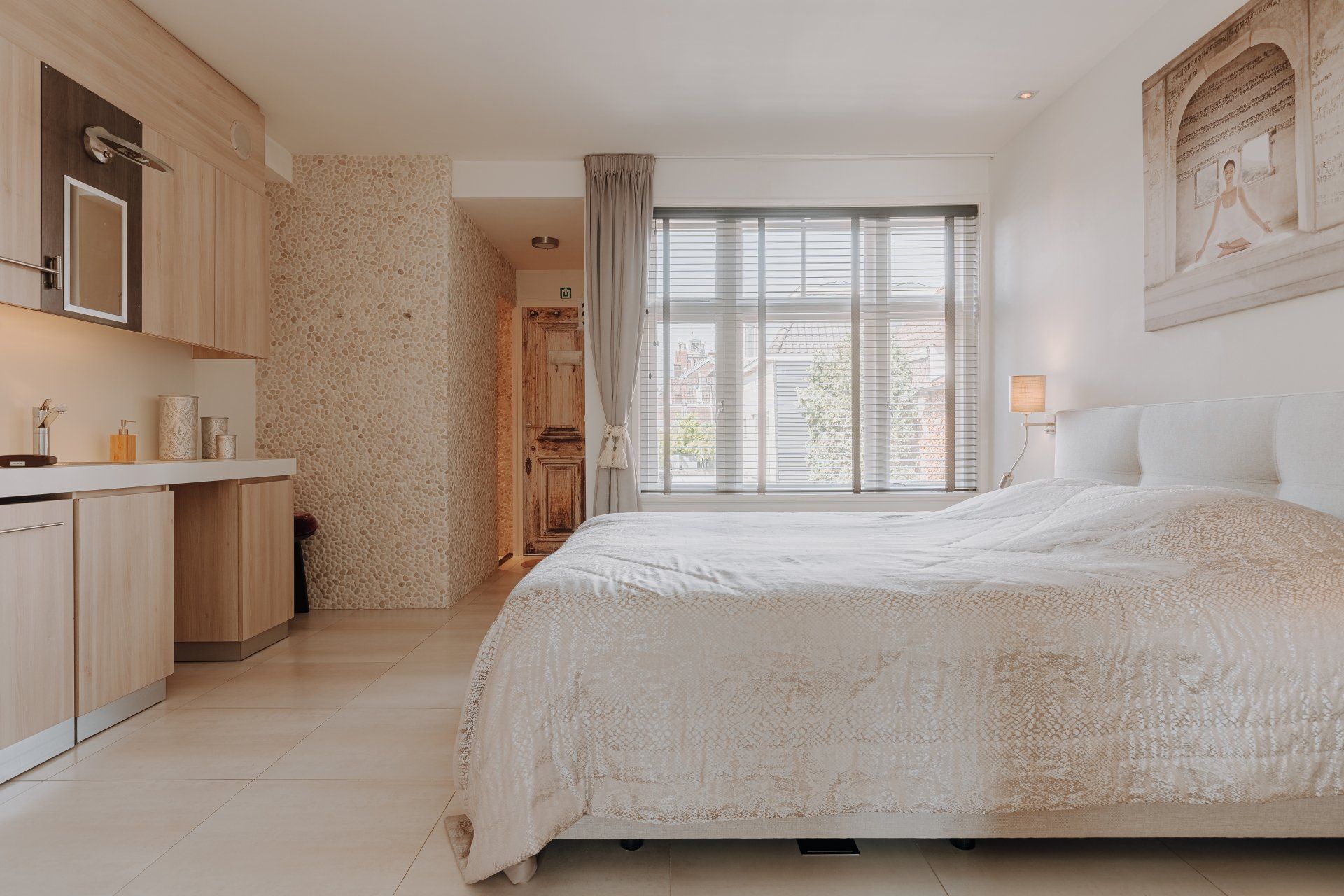 Slaapkamer met een bed, kitchenette en een raam met jaloezieën. Neutrale kleuren en zachte verlichting.