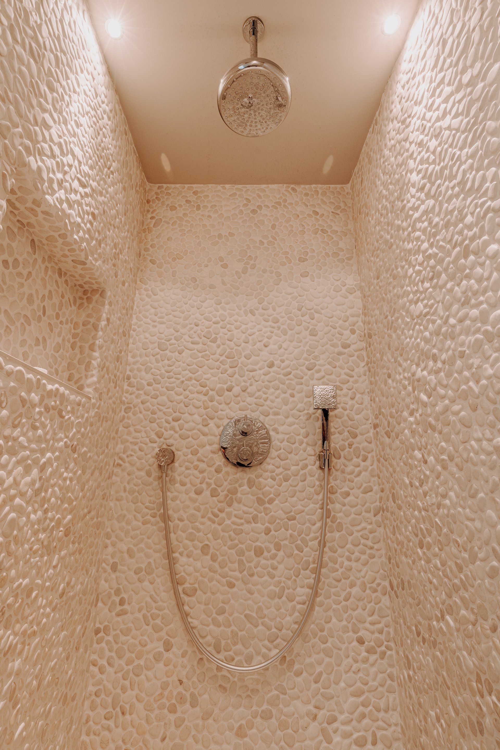 Douchecabine met wanden van gestructureerde kiezeltegels, inbouwverlichting en een zilverkleurige douchekop met handdouche.