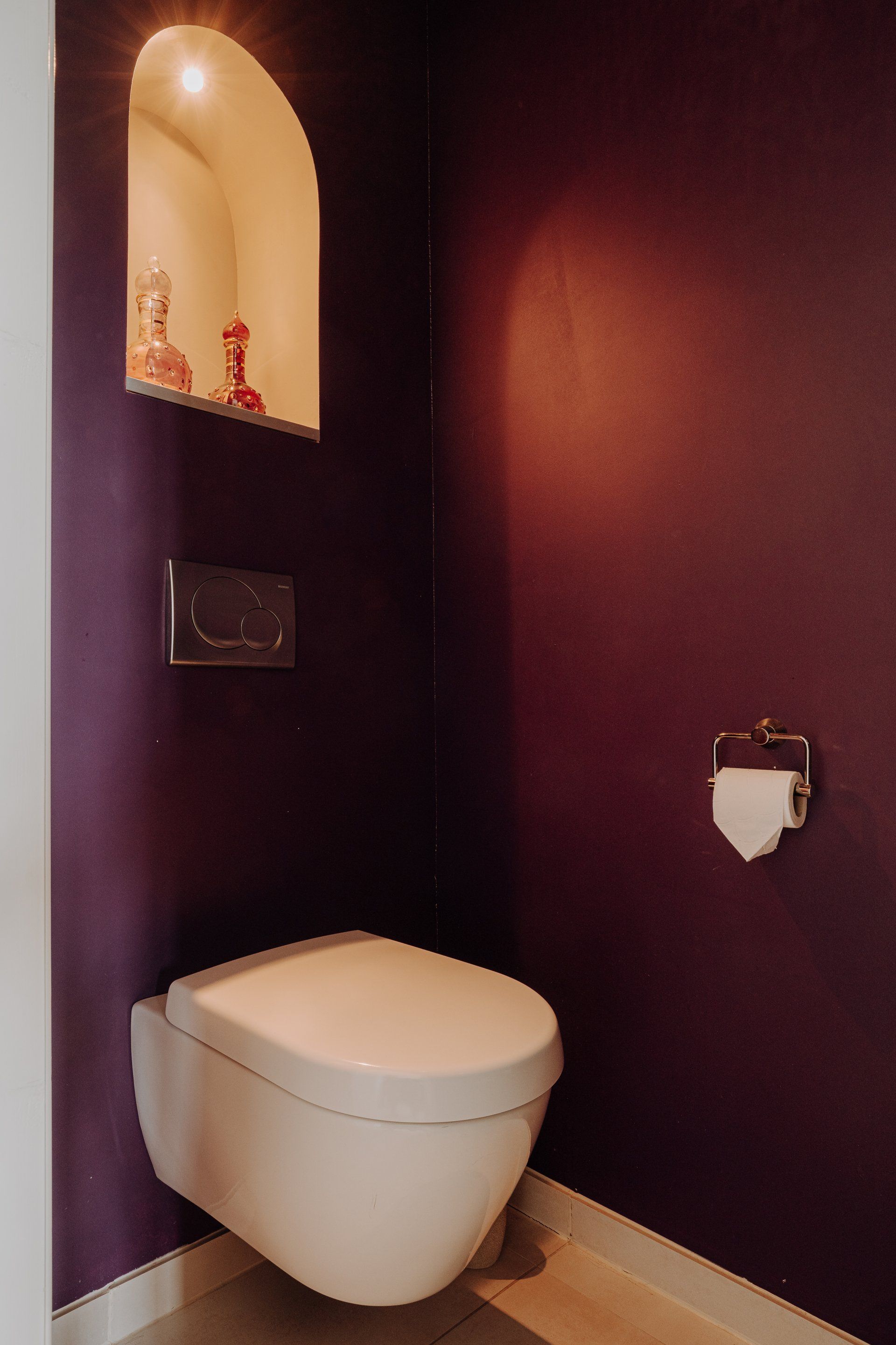 Wandtoilet in een kleine, donkerpaarse wc met een ingebouwde plank waarop decoratieve objecten staan.