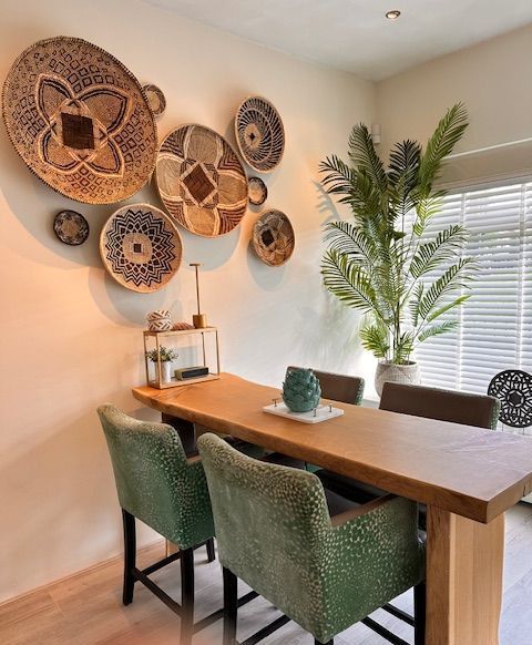 Eetkamer met houten tafel, groene stoelen met patroon, decoratieve wandmanden en een grote potplant.