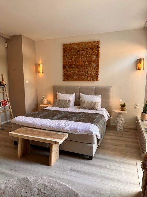 Slaapkamer met een bed, bankje en wanddecoratie, uitgevoerd in neutrale tinten, met houten accenten en wandverlichting.