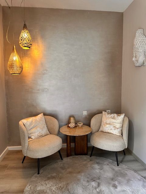 Gezellige zithoek met beige stoelen, een klein houten tafeltje en egyptische hanglampen tegen een bronzen muur met structuur.