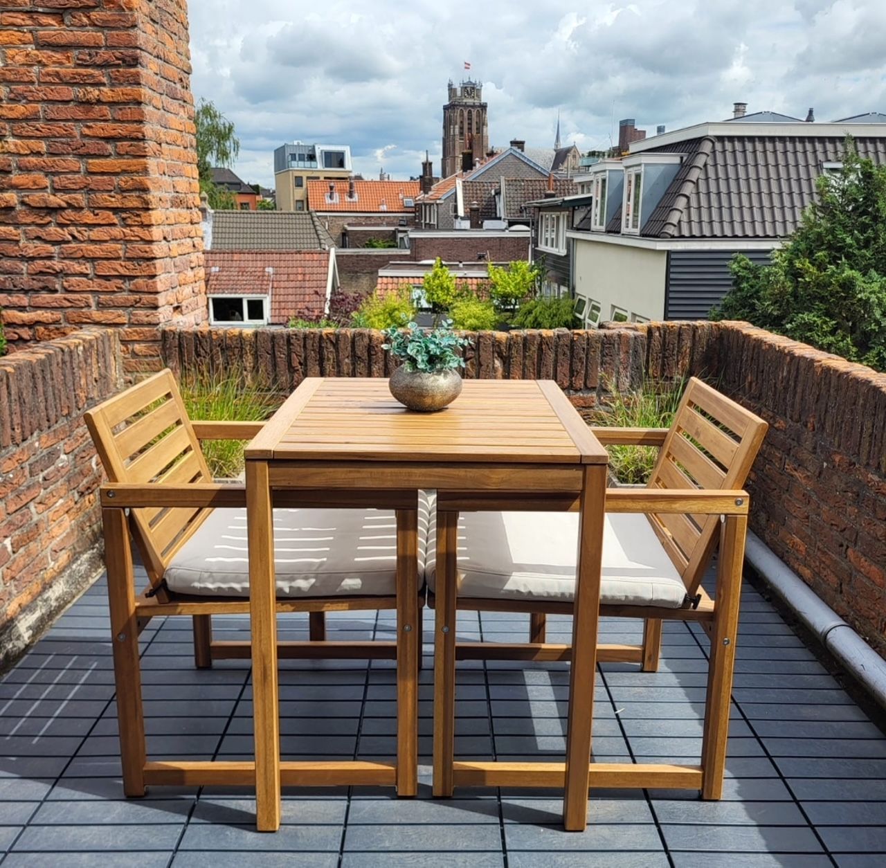Tuinset: vierkante houten tafel, twee stoelen met kussens. Stadsgezicht met gebouwen en kerktoren.