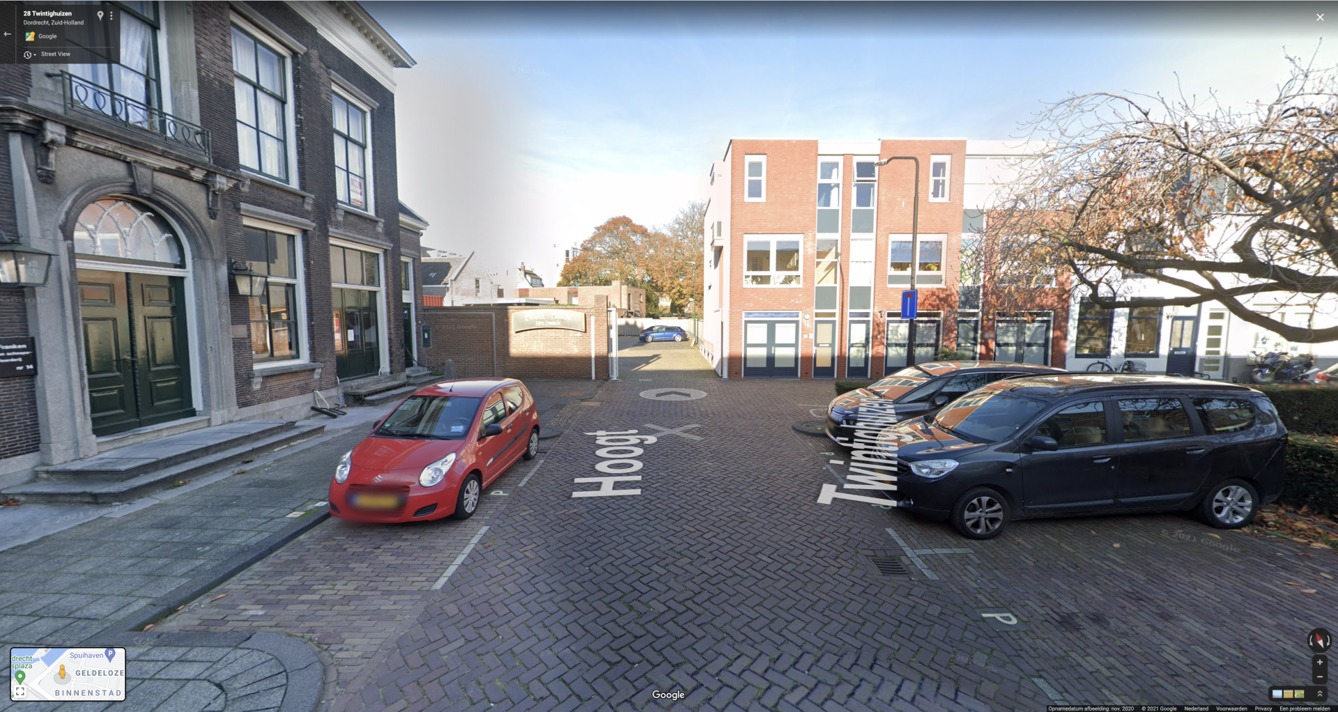 Straatbeeld met auto's geparkeerd bij een bakstenen gebouw en een rode auto links ervan.