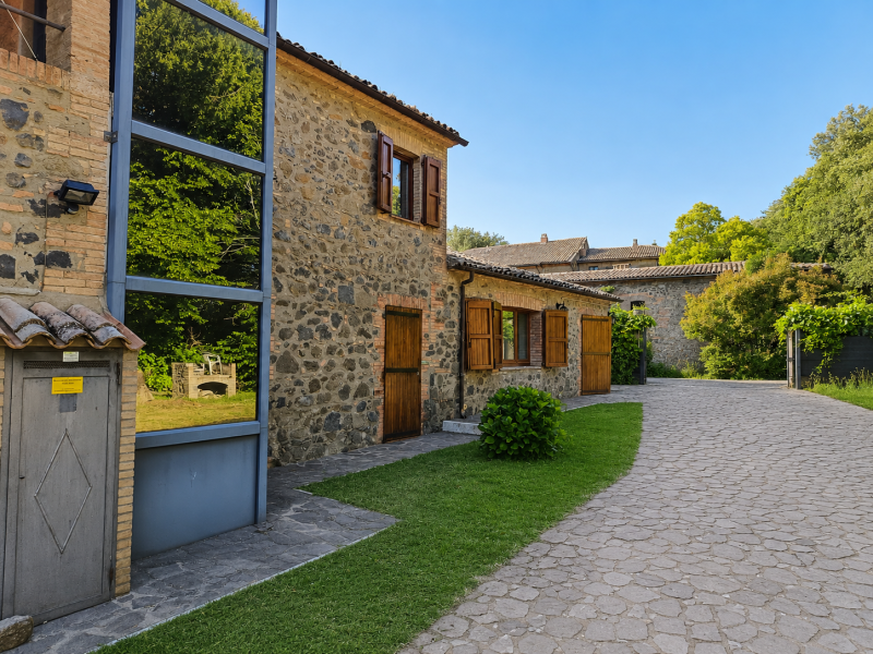 Cortile in pietra con case rustiche, persiane in legno e un sentiero lastricato sotto un cielo azzurro limpido.