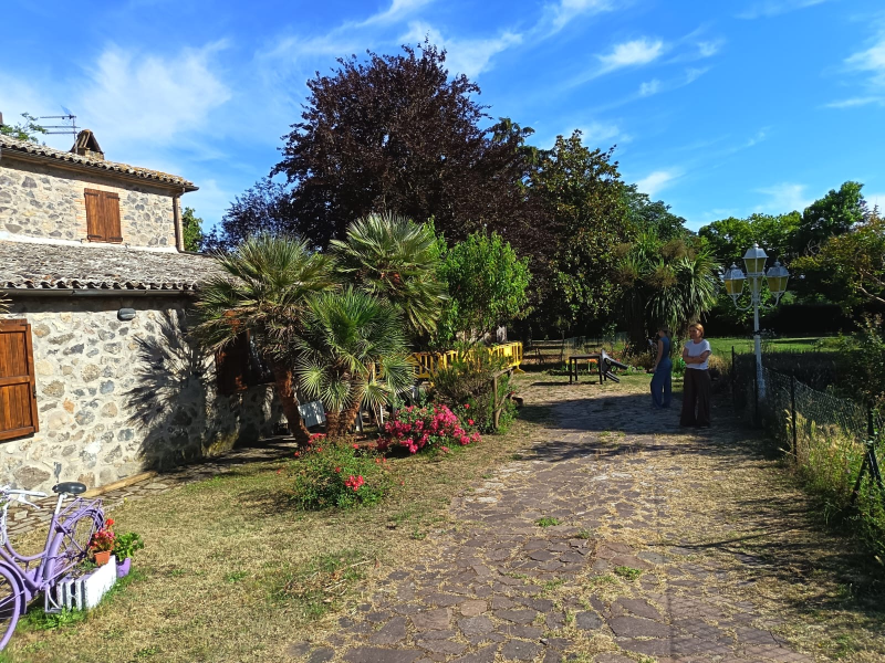 Sentiero nel giardino di una casa in pietra con palme, fiori e due persone in lontananza sotto un cielo azzurro.