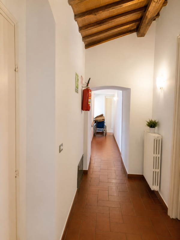 Lungo e stretto corridoio con pavimento in piastrelle di terracotta, pareti bianche, soffitto con travi in ​​legno e una sedia in fondo.