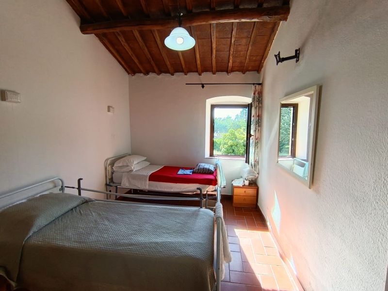 Piccola camera da letto con due letti, pareti bianche, soffitto in legno e luce naturale proveniente da una finestra.