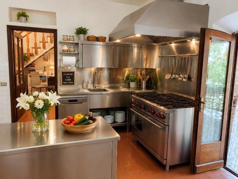 Cucina moderna in acciaio inox con isola, piano cottura, porta a vetri apribile e vaso di fiori sul bancone.