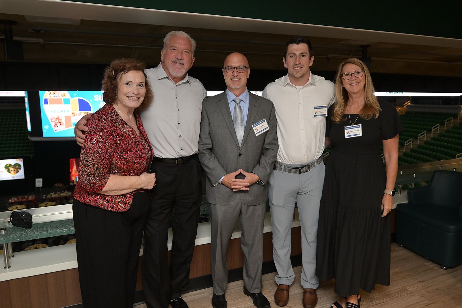 Four adults stand together in a row smiling in what appears to be a stadium luxury suite.