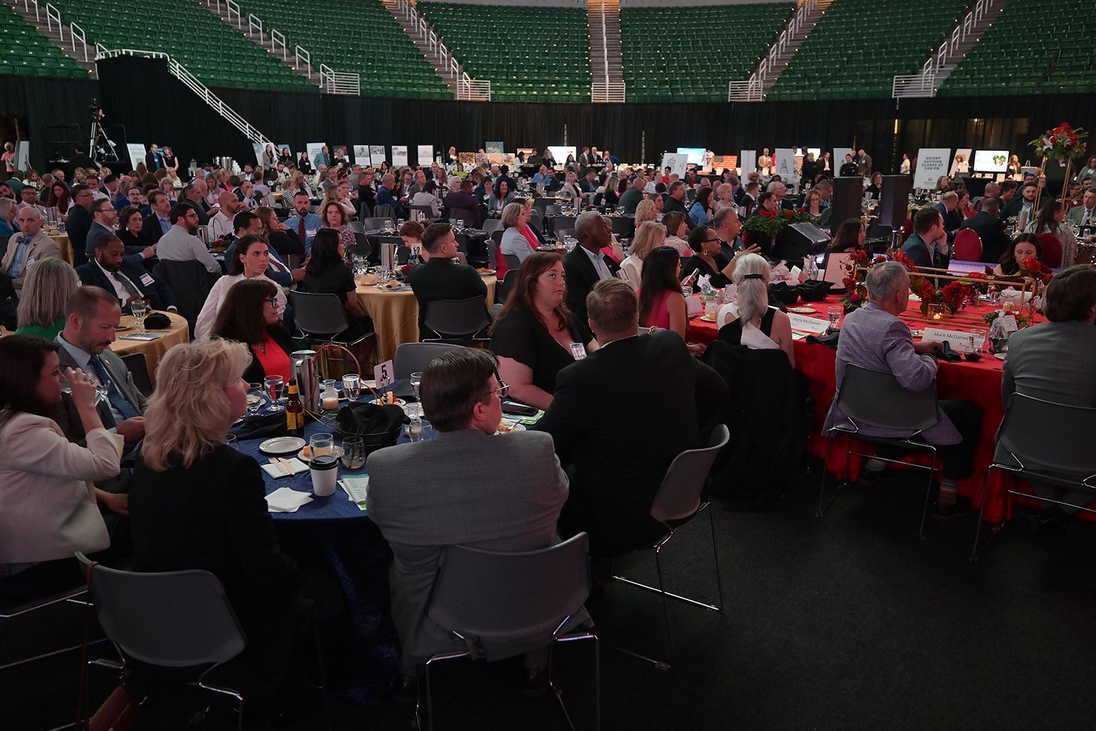 A large, well-lit ballroom filled with people seated at tables for a banquet in front of a green stadium seating area.