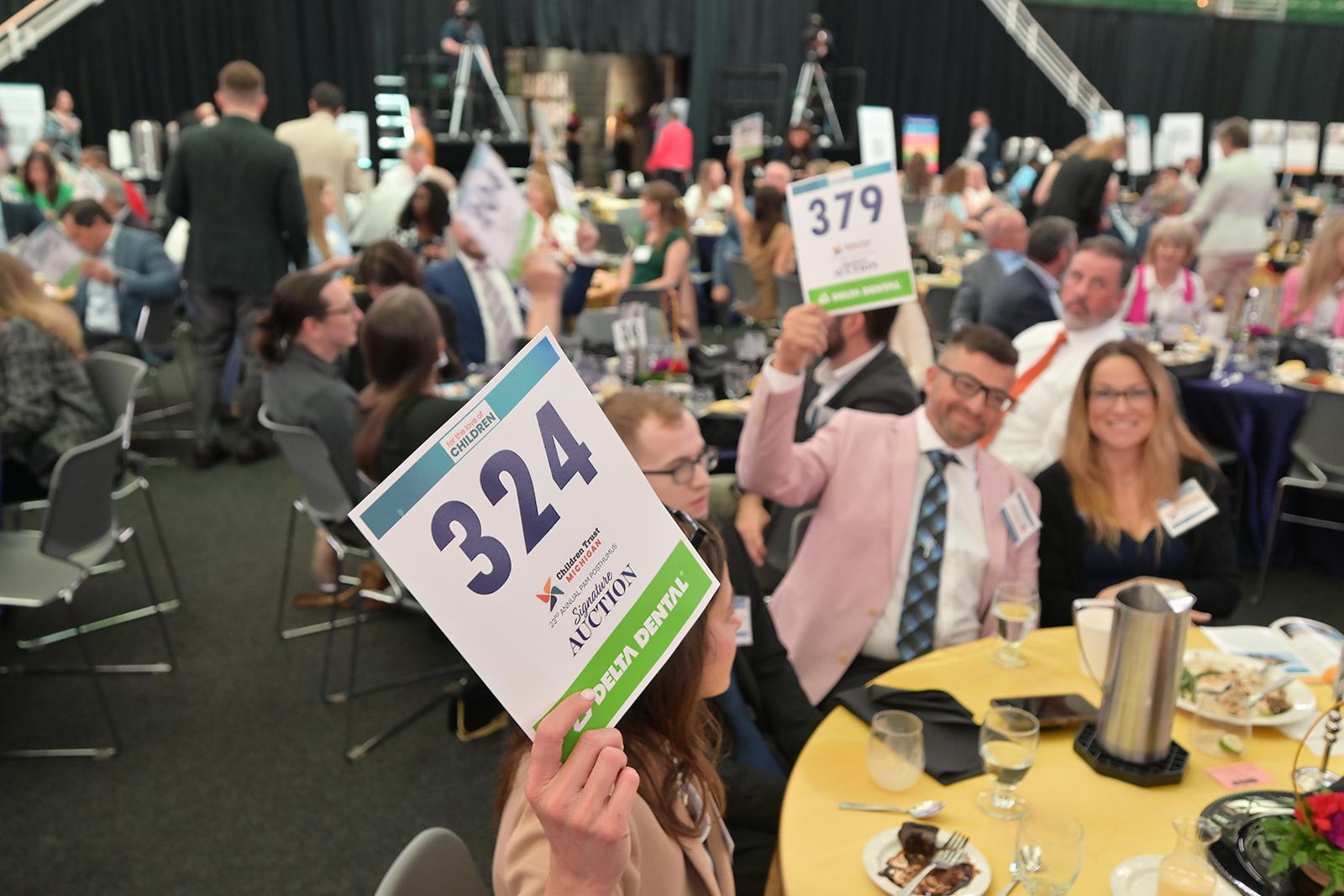 Attendees at an event holding up numbered bidding paddles at a table in a large, crowded hall.