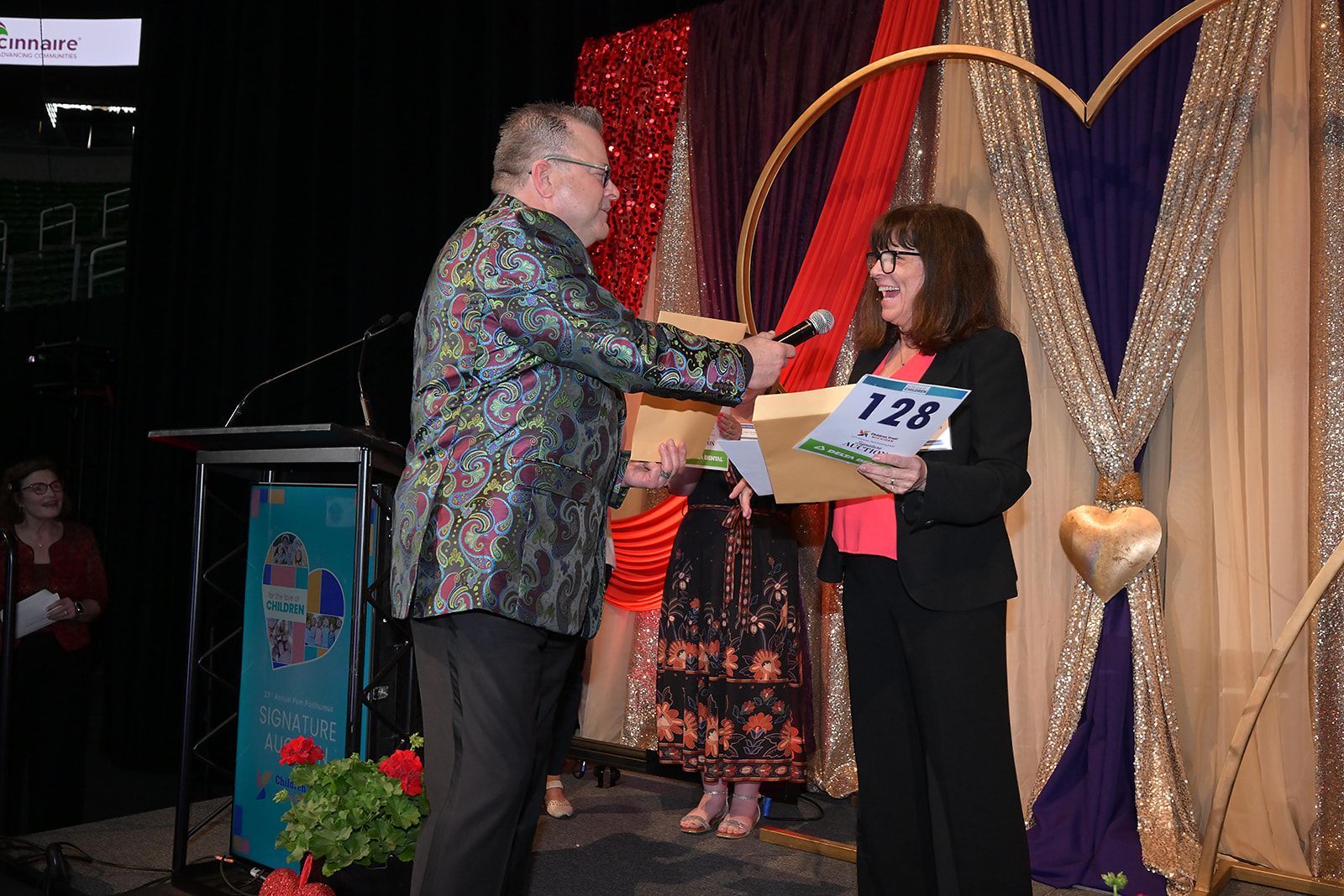 A person in a patterned blazer presents a folder to another person on a stage decorated with purple and gold fabric.
