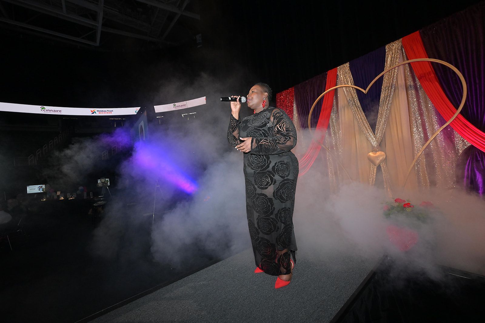A person in a black patterned gown sings into a microphone on a stage filled with theatrical fog, before a heart backdrop.