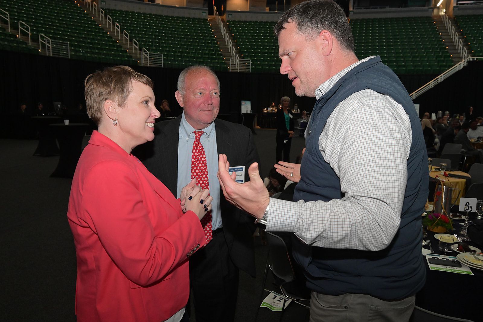 Three people in a large arena interact, with one speaking while the others listen in a professional, social setting.