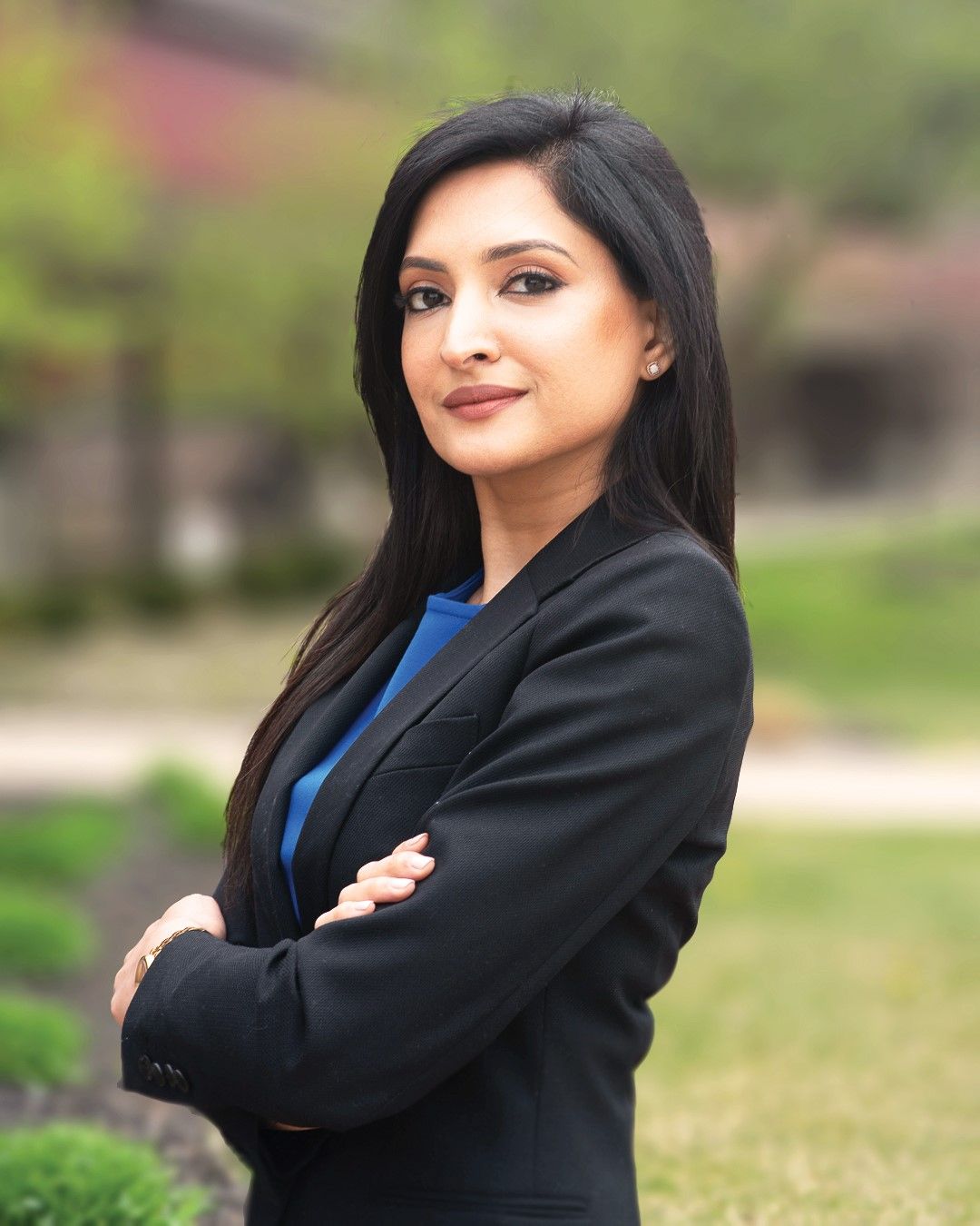 Professional headshot of a person wearing a black blazer and blue top, standing with arms crossed in an outdoor setting.