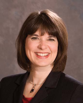 A person with shoulder-length brown hair and a friendly smile, wearing a dark blazer and a pendant necklace.