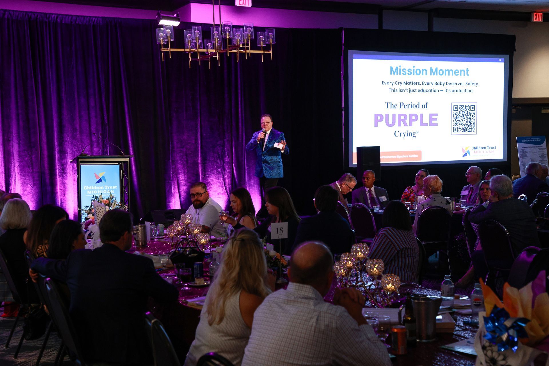 Speaker on stage at a purple-lit banquet, addressing seated guests with presentation slides and a QR code on screen.