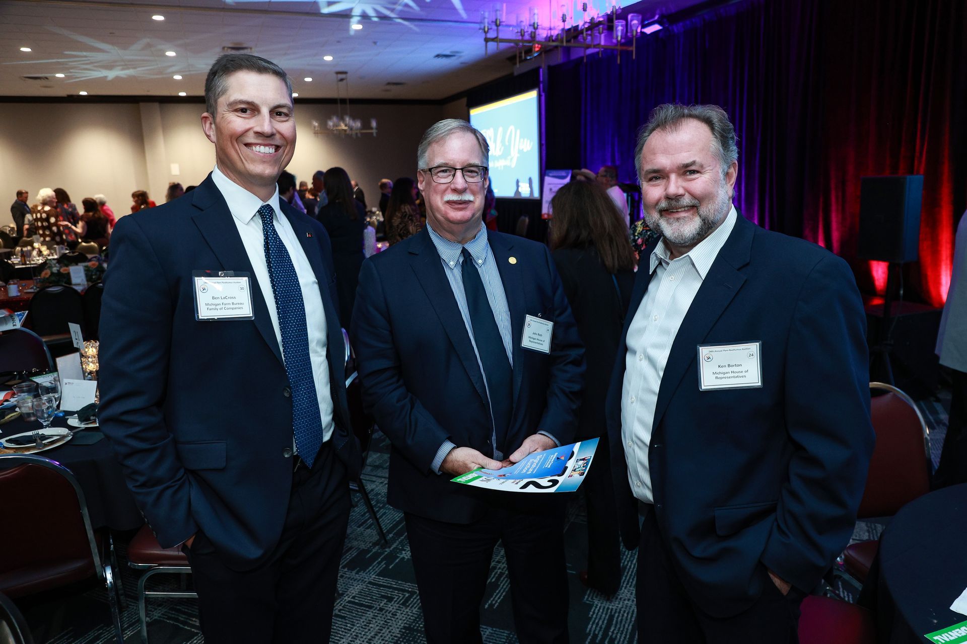 Three men in suits pose at a formal indoor event, with one holding a blue booklet.