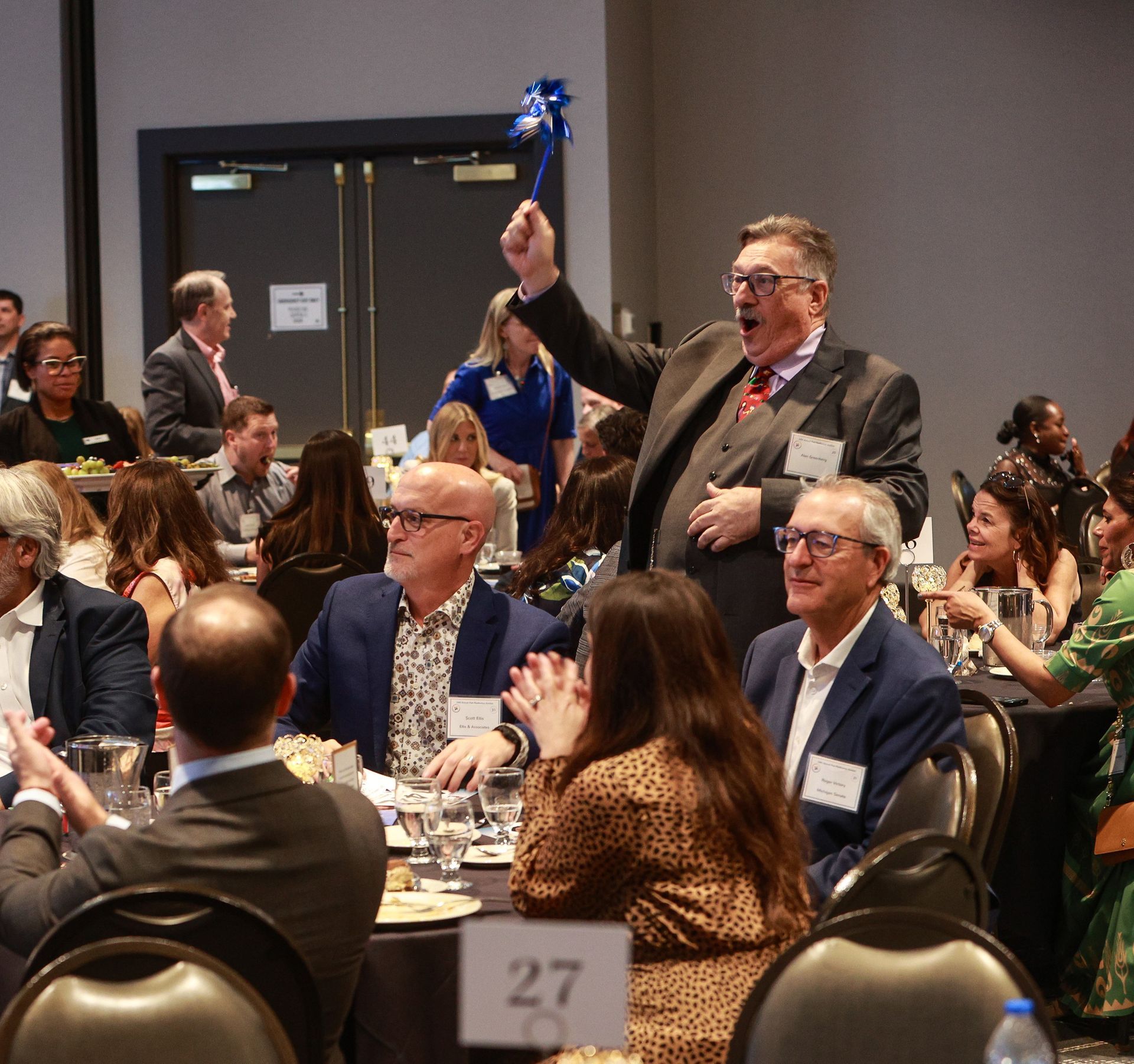 Man in a suit cheering and raising a blue item at a banquet table in a crowded conference hall