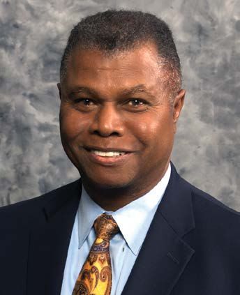 Headshot of a smiling man in a dark blue suit, light blue collared shirt, and a gold-toned patterned tie against a grey.