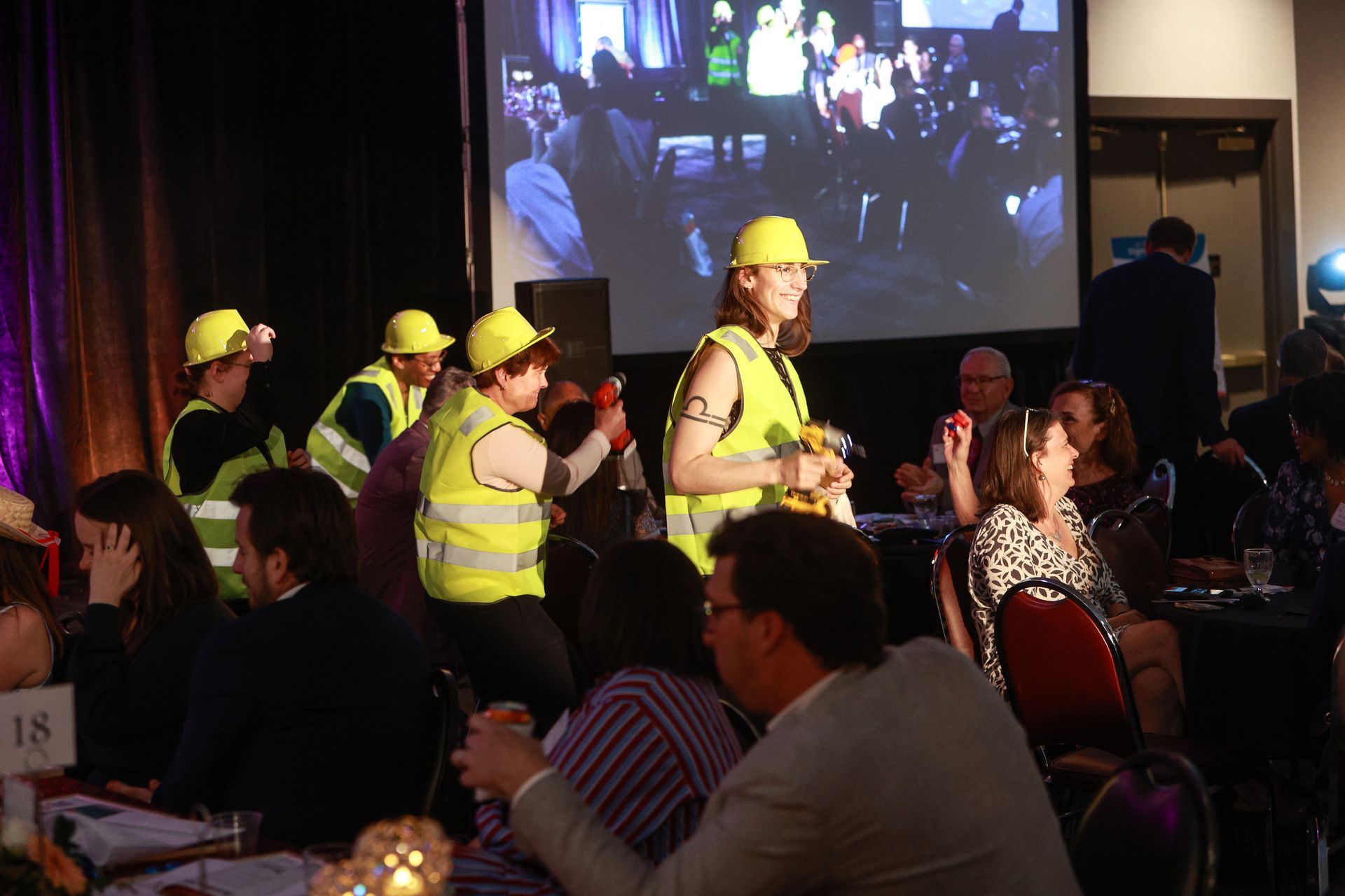 Construction workers in yellow vests and hard hats standing at a formal indoor event with seated guests.
