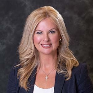 A headshot of a smiling person with long blonde hair, wearing a dark blazer over a white top and a silver cross necklace.