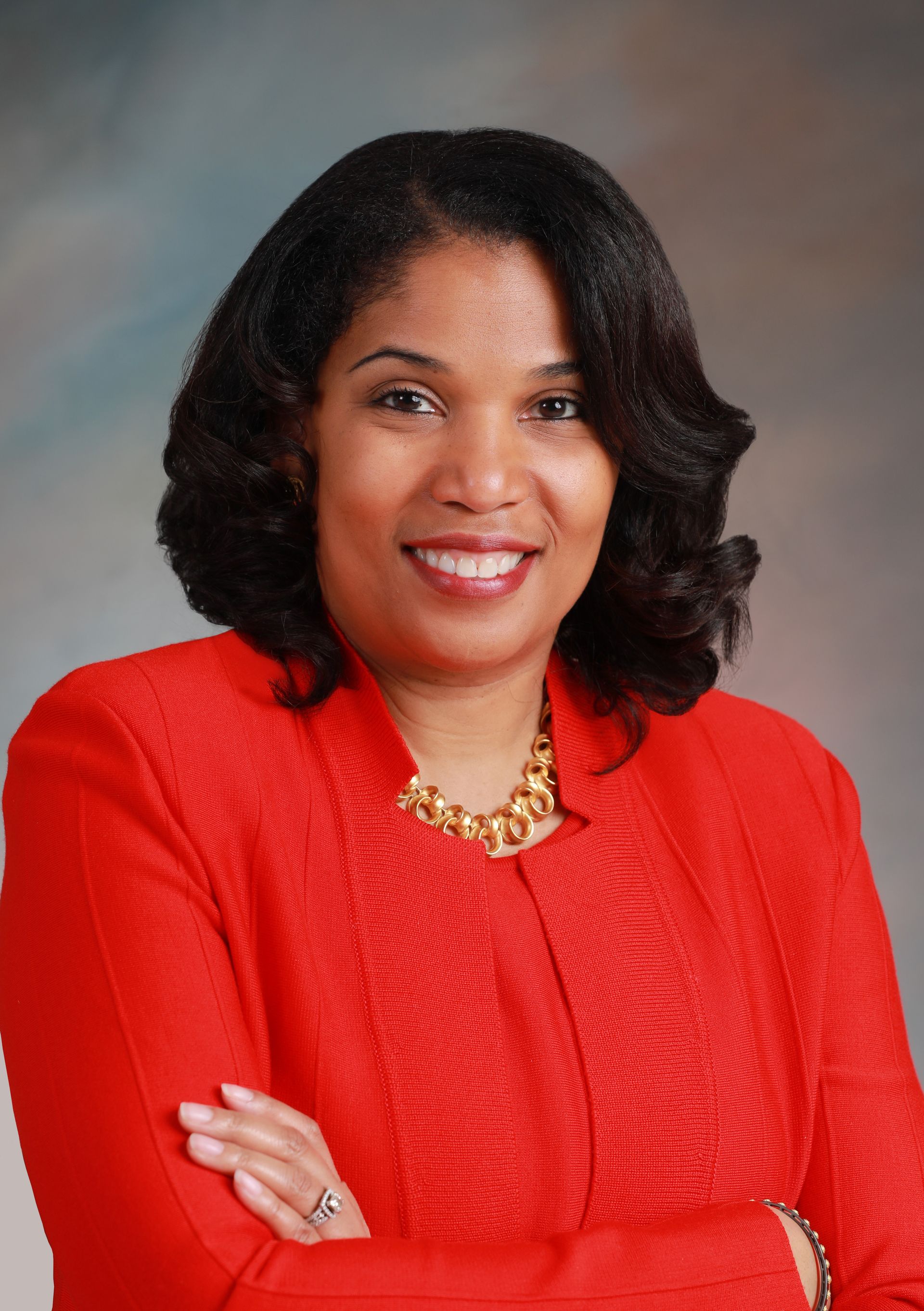 A smiling woman with shoulder-length dark hair wears a bright red blazer and a gold necklace, posing with arms crossed.