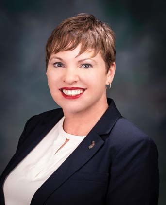 A professional headshot of a person with short hair, smiling, wearing a dark blazer over a white blouse.