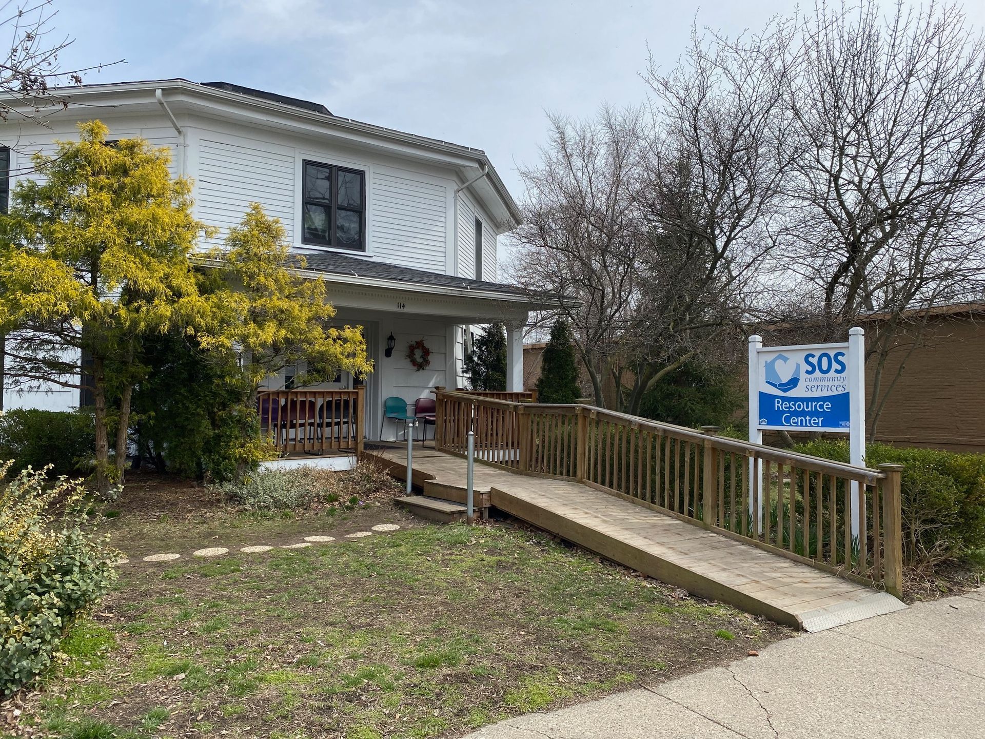 A two-story white house with a wheelchair ramp leading to the front porch and a blue sign for the SOS Community Center.