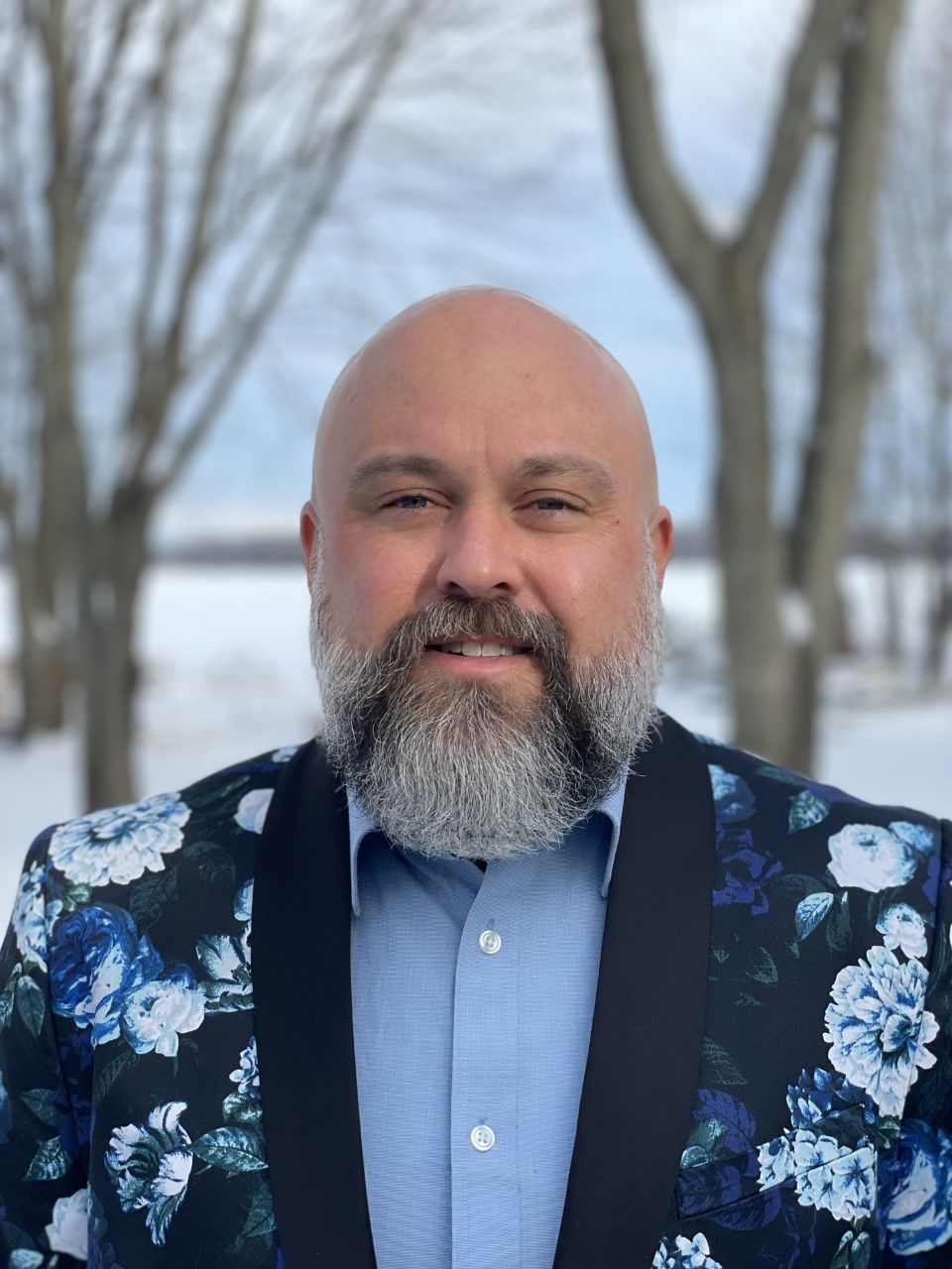 A bald person with a beard wears a blue dress shirt under a black blazer featuring a bold blue and white floral pattern.