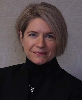 A person with short, light hair wearing a black turtleneck and blazer against a light, patterned background.
