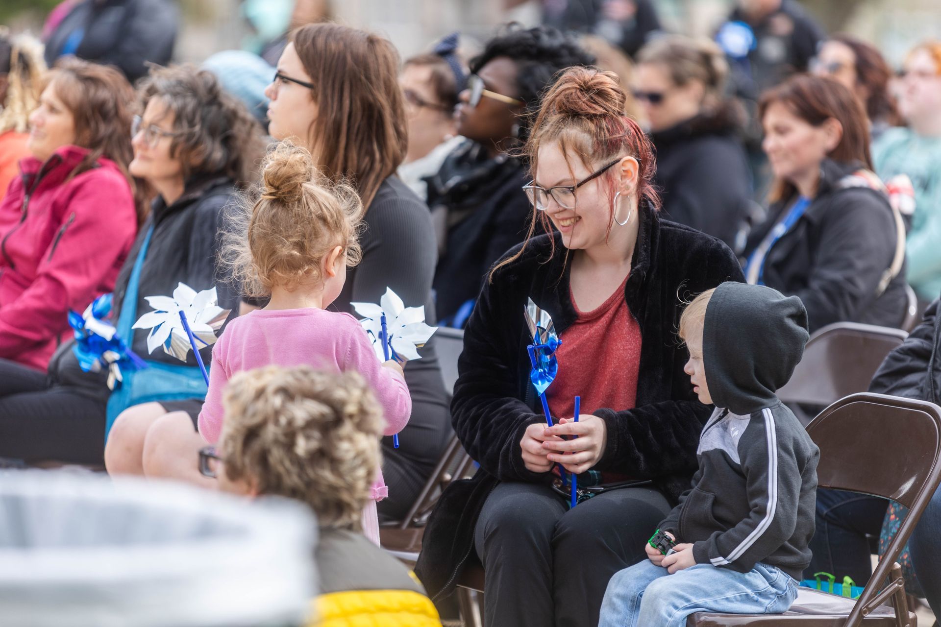 People seated outdoors hold blue pinwheels at an event, with children among the group smiling and looking around.