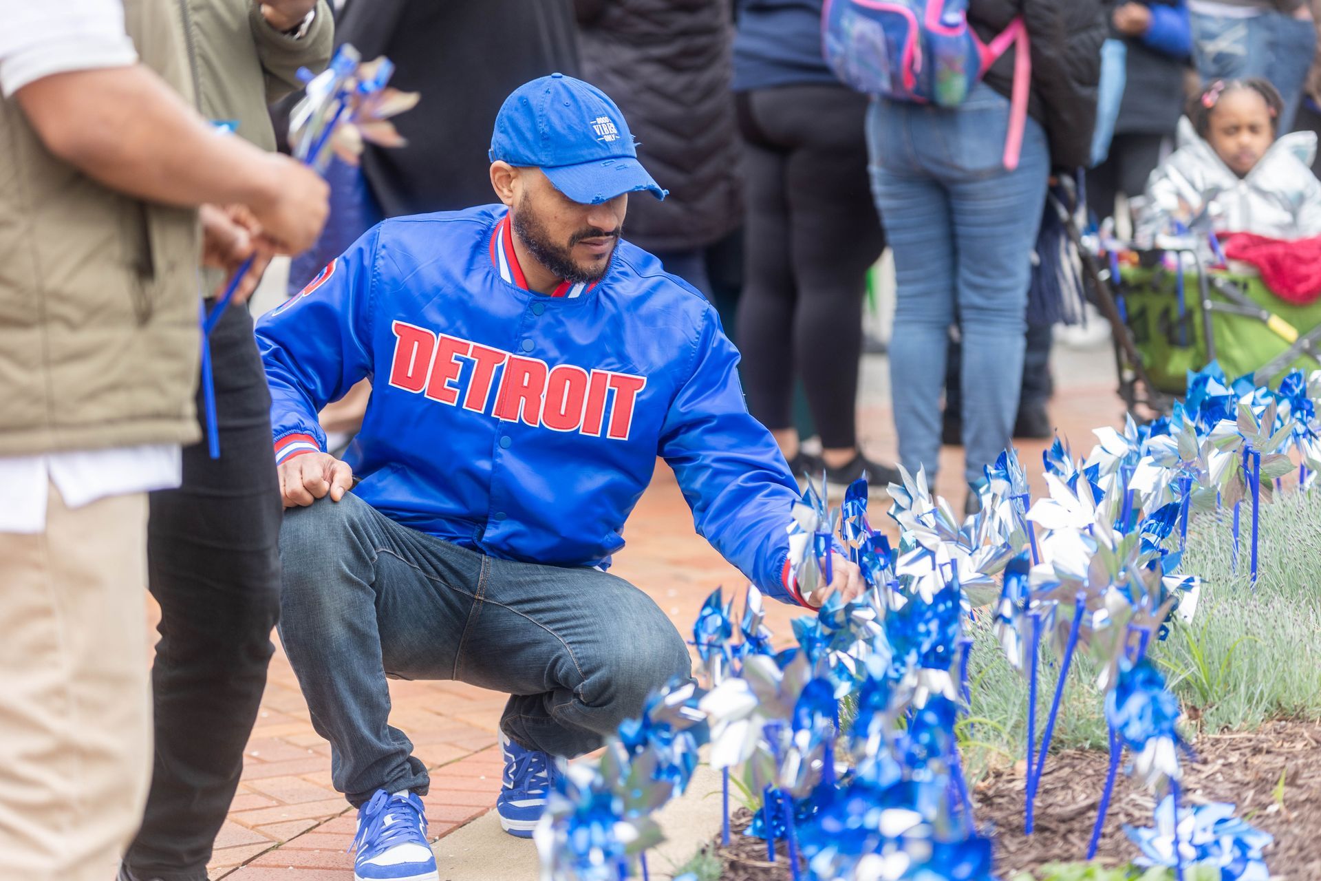 A person in a blue Detroit jacket and cap crouches to place a small blue pinwheel in a garden bed with many others.