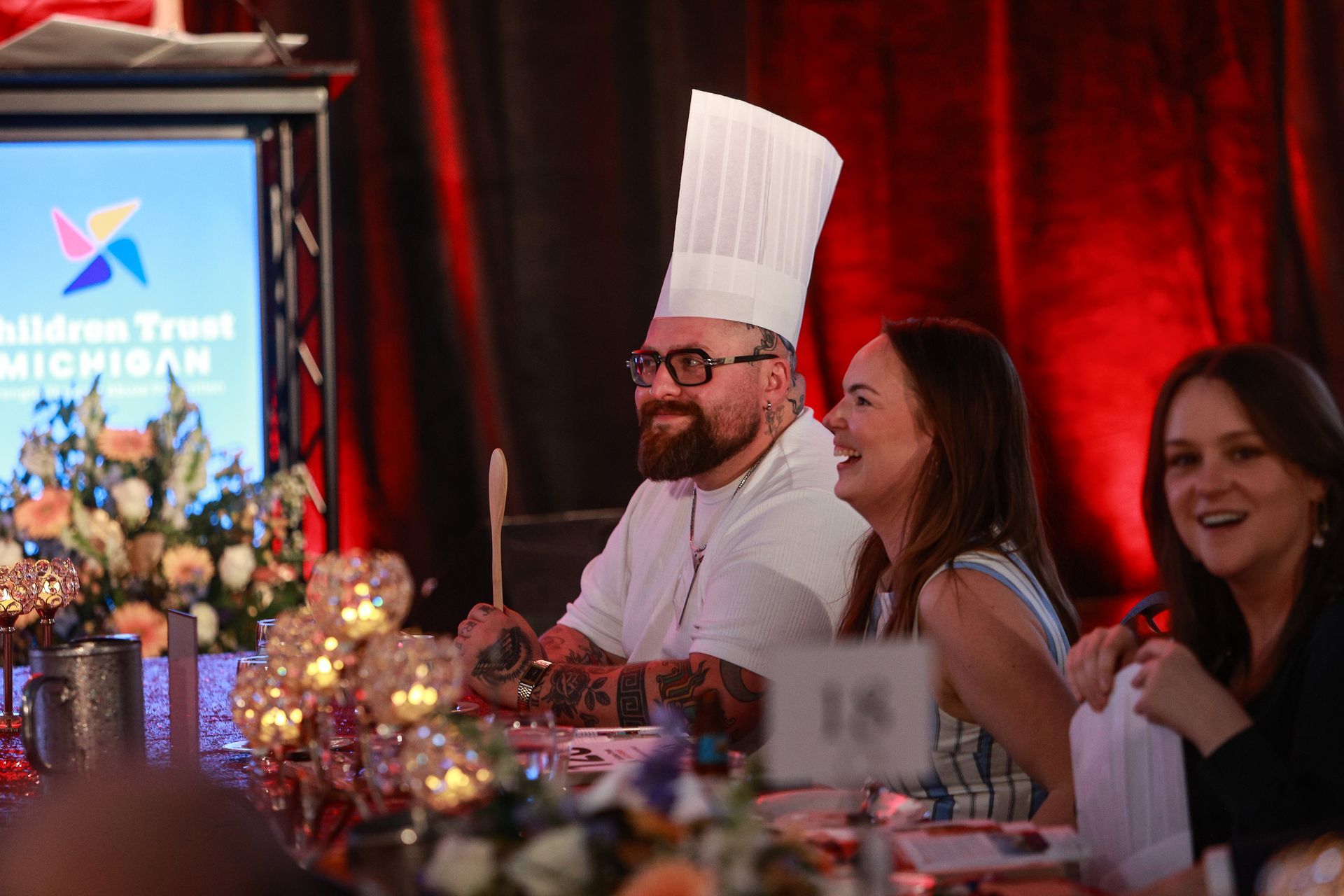 Chef in white hat smiles at banquet table beside seated guests, with floral centerpiece and lit stage behind them