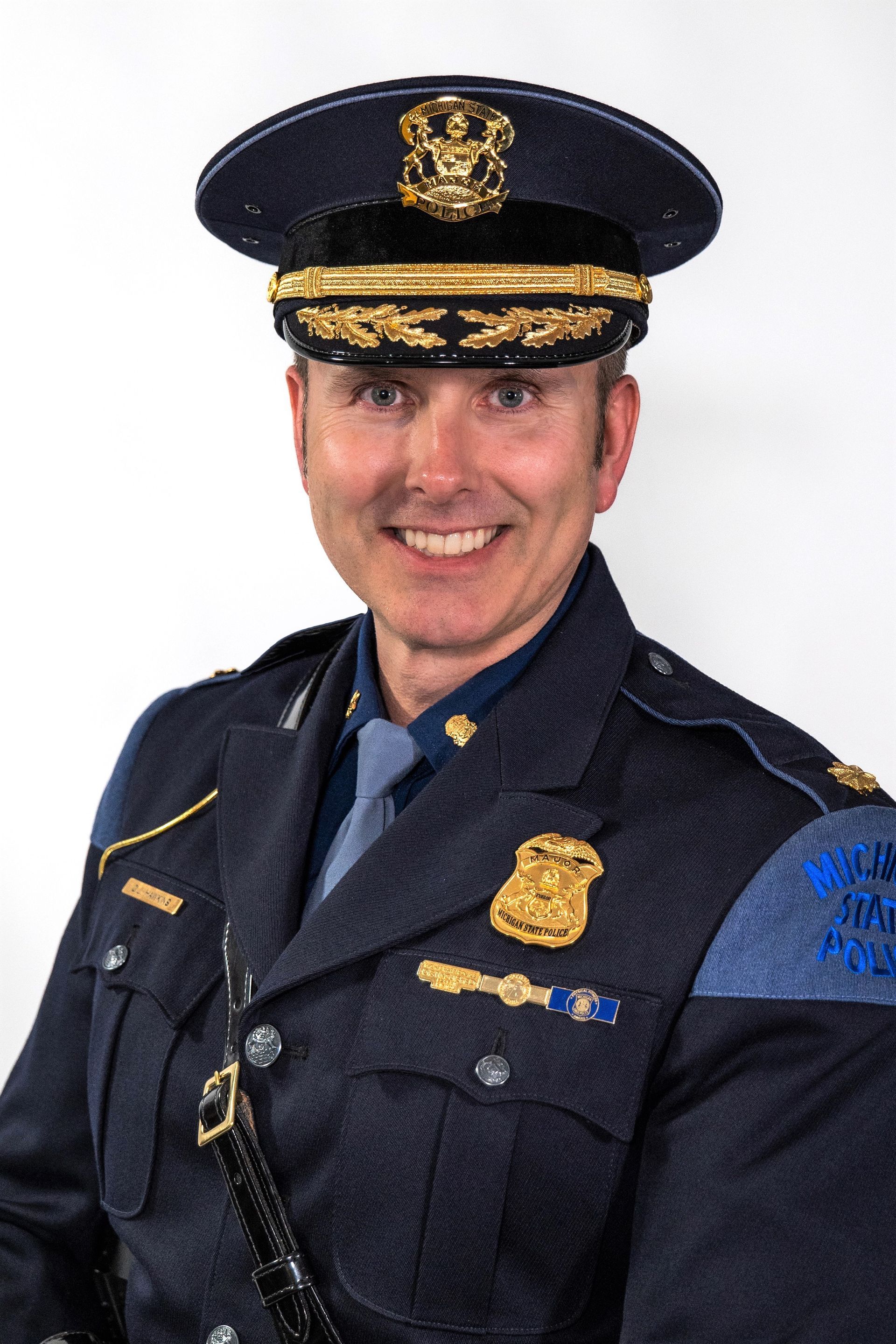 A person in a dark blue official police uniform with a peaked hat and badge, smiling against a white background.