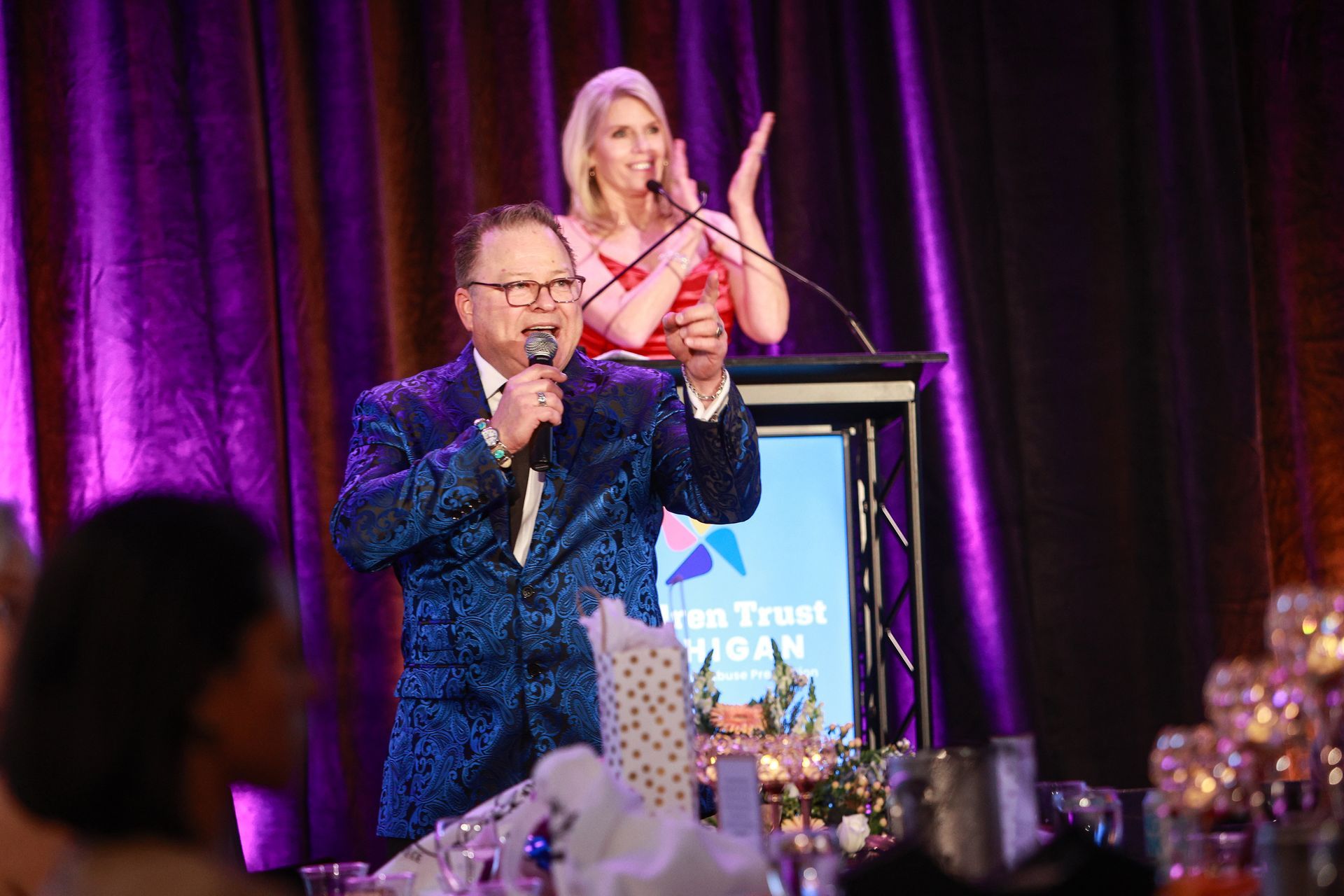 Two speakers onstage at a podium, one holding a microphone, against purple lighting at a gala event.