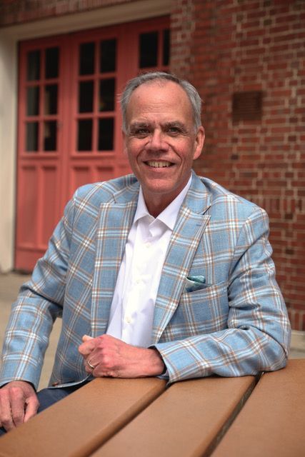 A person wearing a light blue plaid blazer and white collared shirt smiles while sitting at a table near a brick building.