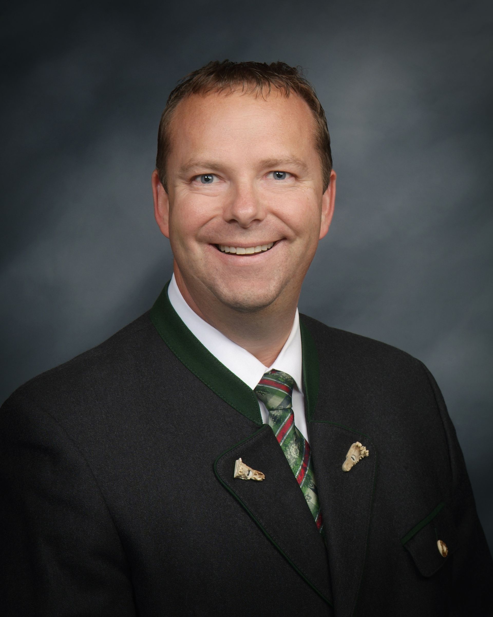 A person wearing a dark suit with green trim and a patterned tie smiles against a grey background.