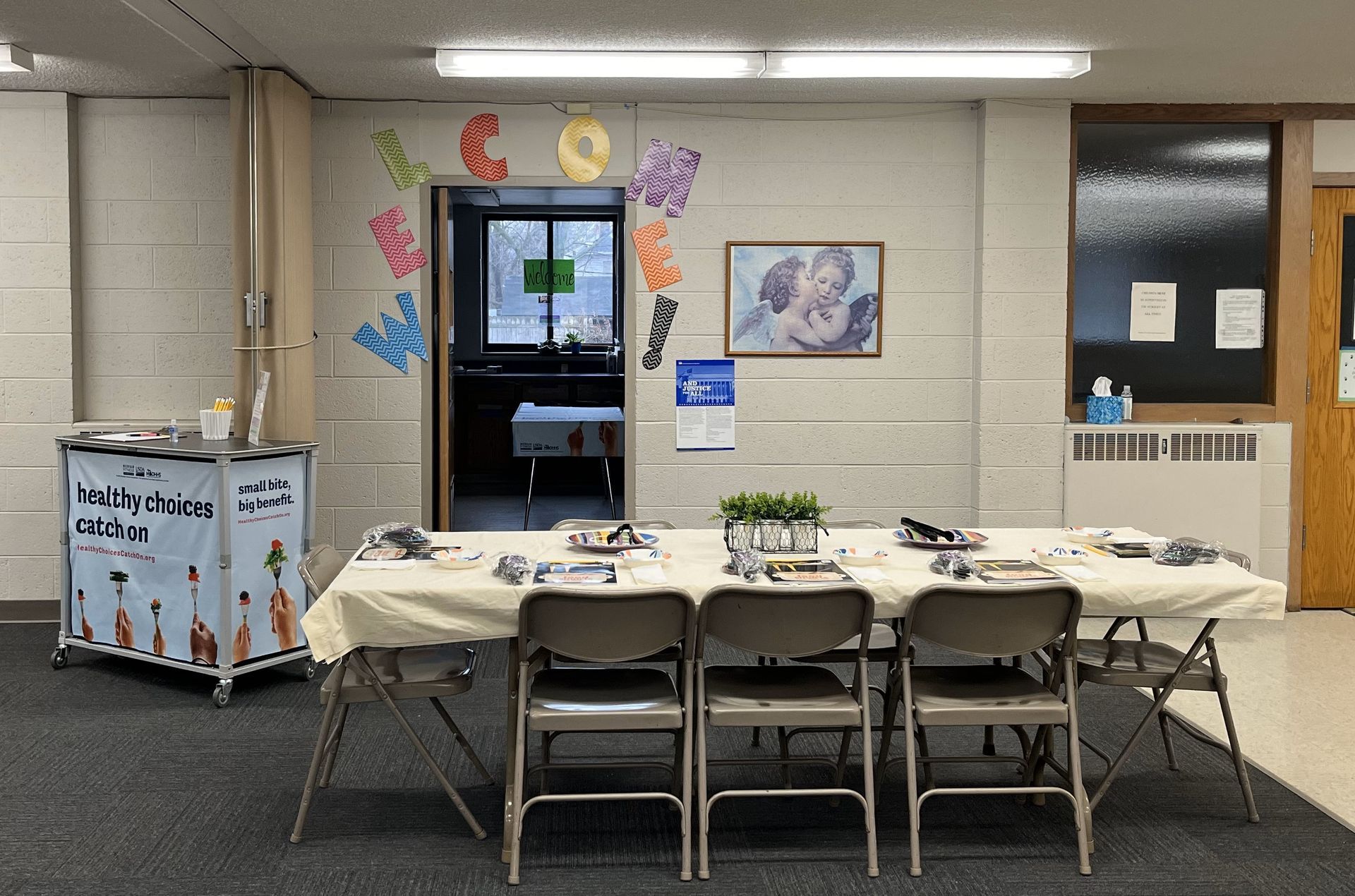 A table set for a small group meeting in a room with a doorway, decorations, and a rolling cart on the left.