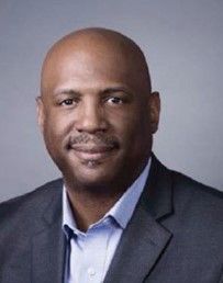 A professional headshot of a person wearing a dark grey blazer and a light blue collared shirt against a grey background.