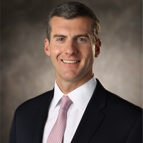 A man in a suit and light pink tie smiles against a neutral gray background.