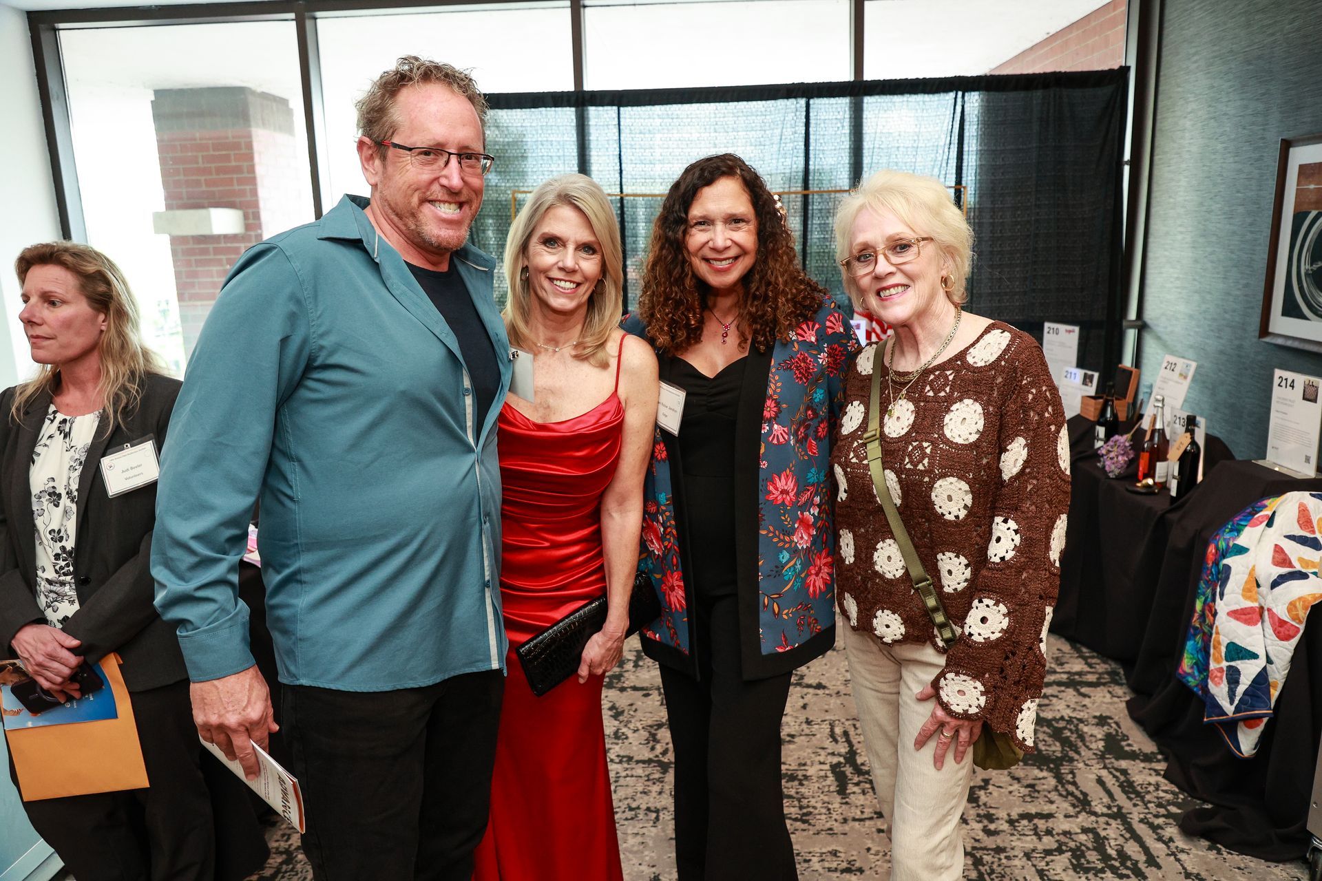 Four people pose indoors at an event, including one man in a teal shirt and two women in red and black dresses.